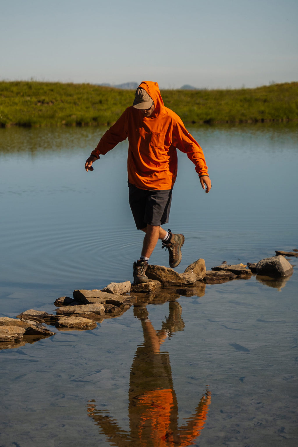 Wearing the 686 Men's Everywhere® Hybrid Short - Relaxed Fit, a person in an orange hoodie carefully crosses stepping stones on a calm lake, with their reflection mirrored in the water against a grassy field and blue sky.