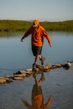Wearing the 686 Men's Everywhere® Hybrid Short - Relaxed Fit, a person in an orange hoodie carefully crosses stepping stones on a calm lake, with their reflection mirrored in the water against a grassy field and blue sky.