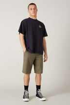 A young man with a neutral expression stands against a plain background, wearing a loose black T-shirt and 686 Men's Everywhere® Hybrid Short - Relaxed Fit in olive green, paired with black crew socks and high-top sneakers, hands in his pockets.