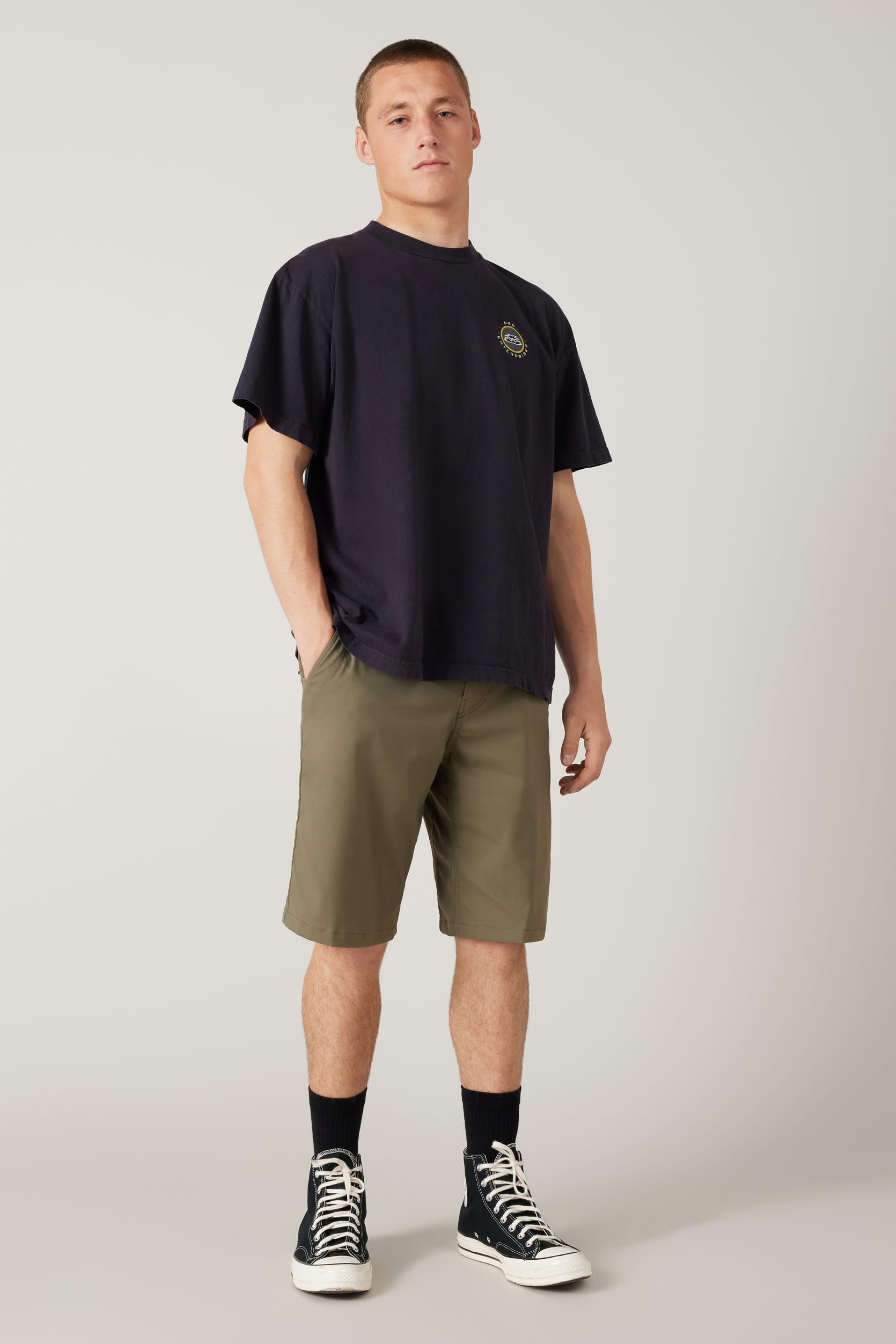 A young man with a neutral expression stands against a plain background, wearing a loose black T-shirt and 686 Men's Everywhere® Hybrid Short - Relaxed Fit in olive green, paired with black crew socks and high-top sneakers, hands in his pockets.