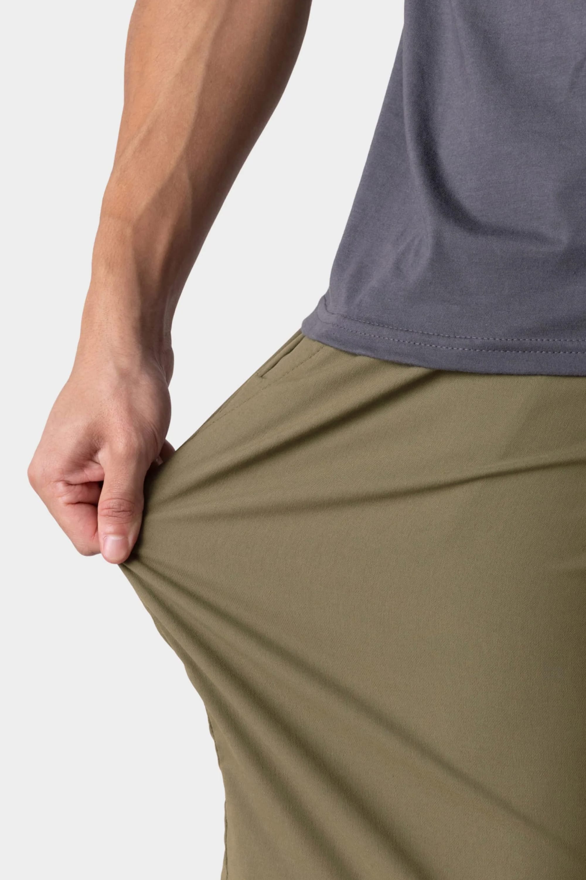 A person wearing a gray shirt and 686 Men's Everywhere® Hybrid Short - Relaxed Fit in olive green stretches the fabric outward with one hand, highlighting the flexibility of the 686 shorts.