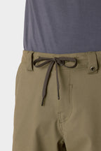 A close-up shows a person wearing 686 Men's Everywhere® Hybrid Short - Relaxed Fit in olive green with a black-and-white drawstring, paired with a tucked-in grayish-blue shirt.