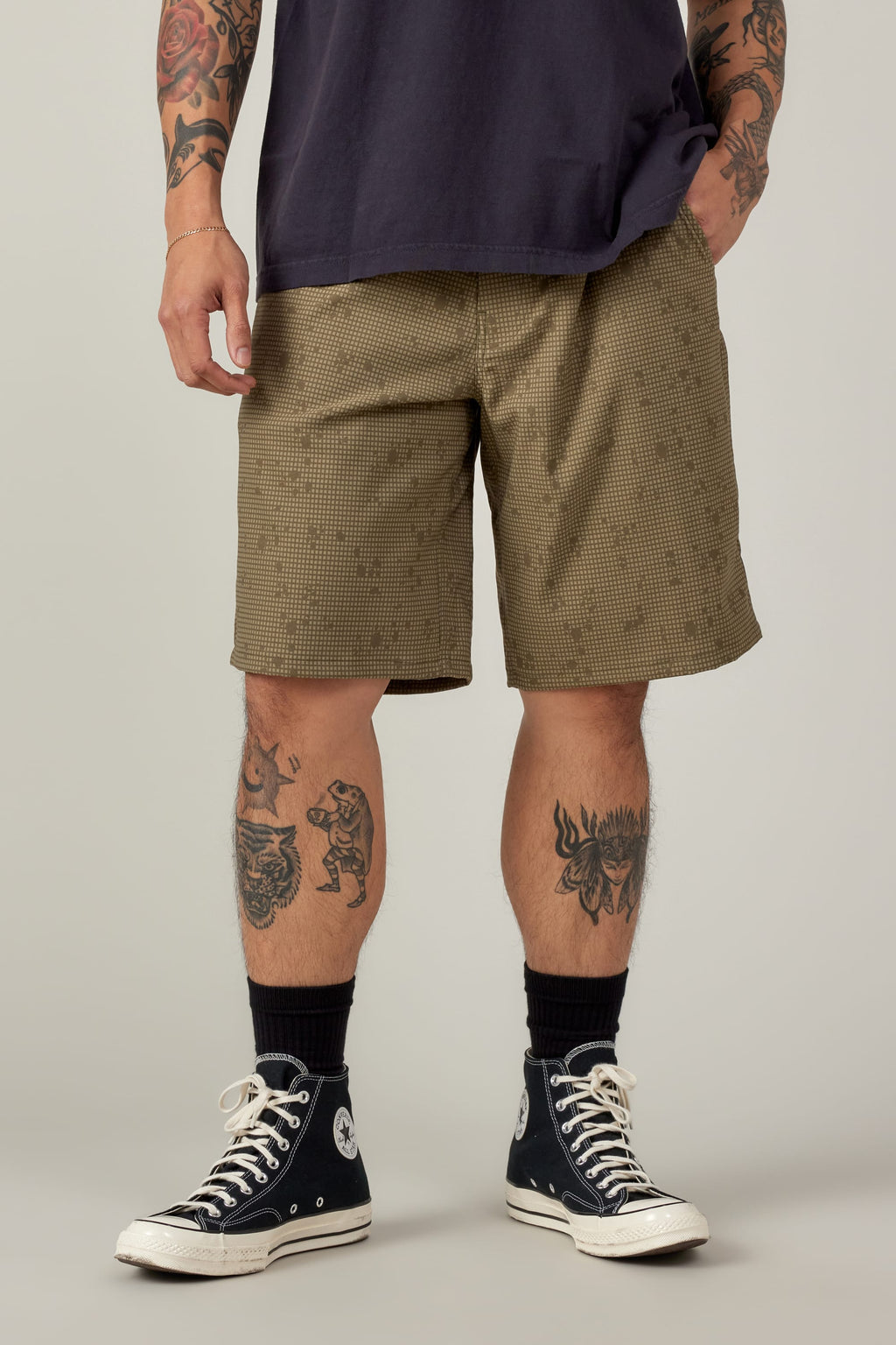 Wearing a dark t-shirt and 686 Men's Everywhere® Hybrid Short - Relaxed Fit in a checked khaki pattern, paired with black socks and Converse. Tattoos cover their arms and legs; image is cropped at shoulders, showing only lower torso and legs.