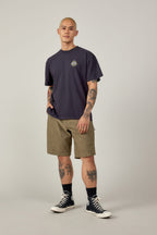 A person with short hair and tattoos wears a dark navy t-shirt, black socks, black high-top sneakers, and 686 Men’s Everywhere® Hybrid Short - Relaxed Fit by 686 in olive green, standing against a plain light gray background.