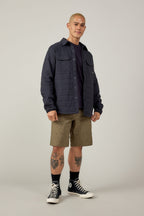 A man with short hair wears a dark quilted jacket over a black shirt, black socks, black Converse sneakers, and 686 Men's Everywhere® Hybrid Short - Relaxed Fit. He stands confidently, showing tattooed legs, against a plain light background.
