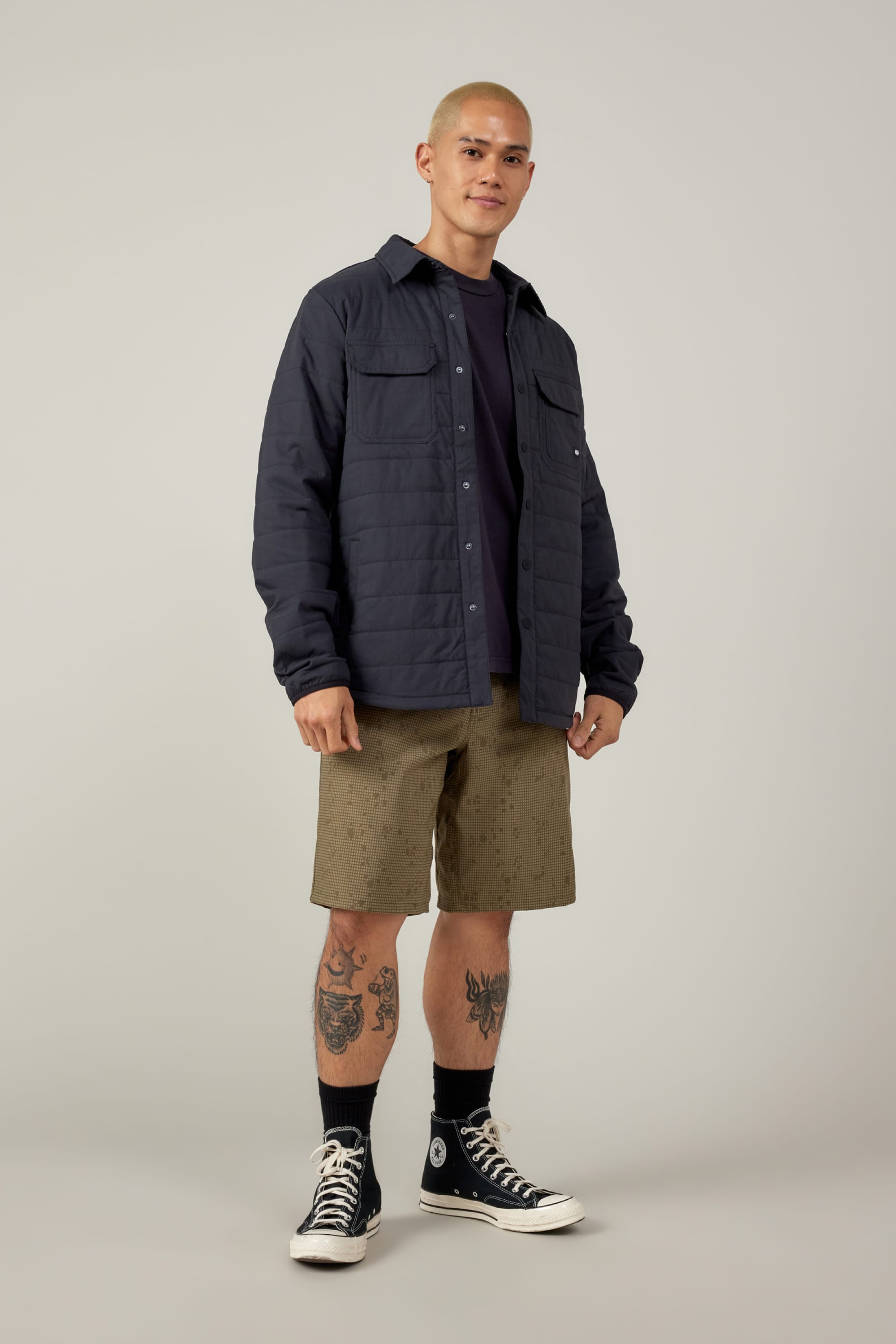 A man with short hair wears a dark quilted jacket over a black shirt, black socks, black Converse sneakers, and 686 Men's Everywhere® Hybrid Short - Relaxed Fit. He stands confidently, showing tattooed legs, against a plain light background.