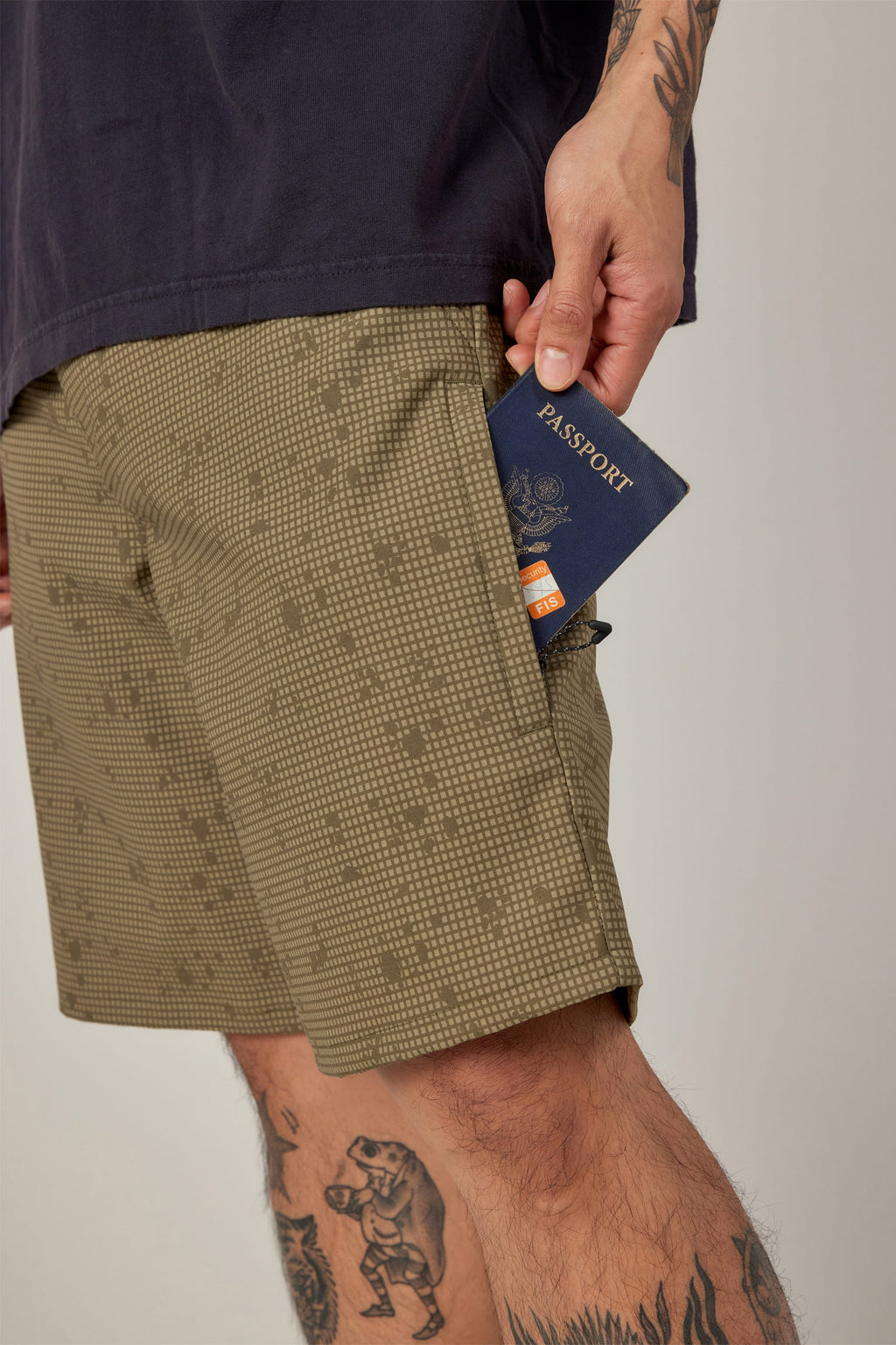 Wearing 686 Men's Everywhere® Hybrid Short - Relaxed Fit in a patterned khaki, a tattooed individual with a bear leg tattoo slips a navy passport into the pocket, beneath a dark shirt. Brand: 686.