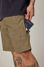 Wearing 686 Men's Everywhere® Hybrid Short - Relaxed Fit in a patterned khaki, a tattooed individual with a bear leg tattoo slips a navy passport into the pocket, beneath a dark shirt. Brand: 686.