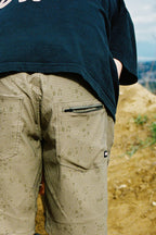 A close-up of a person’s lower back and legs showcases the 686 Men's Everywhere® Hybrid Short - Relaxed Fit in green, paired with a black shirt, standing outdoors on a dirt path with a blurred natural background.