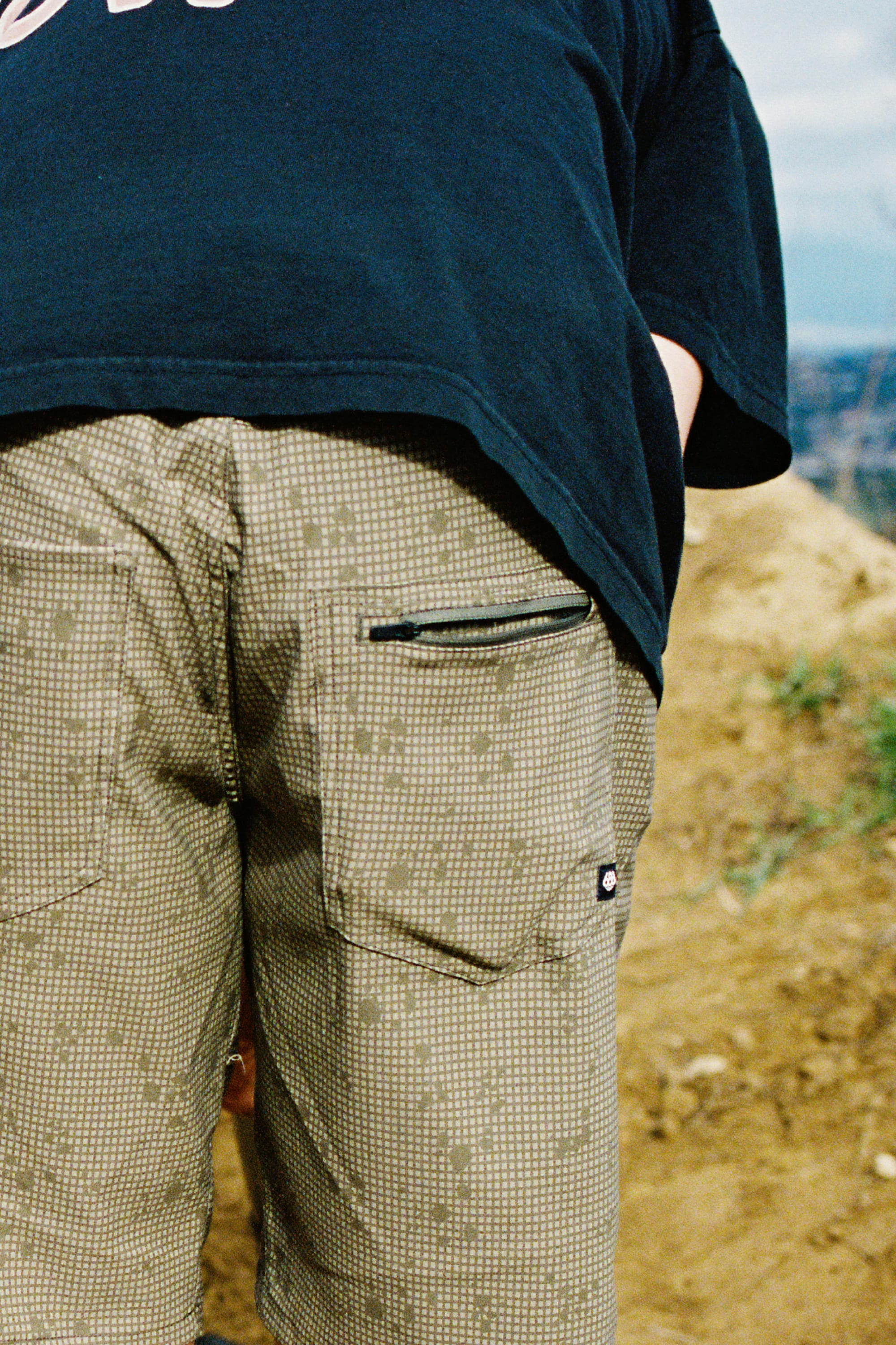 A close-up of a person’s lower back and legs showcases the 686 Men's Everywhere® Hybrid Short - Relaxed Fit in green, paired with a black shirt, standing outdoors on a dirt path with a blurred natural background.