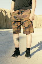 Outdoors on sandy ground, a person wears 686 Men's Everywhere® Hybrid Short - Relaxed Fit with a camo print, a black shirt, black socks, and shoes. A colorful bird tattoo shows on their lower right leg, rocky cliffs in the background.