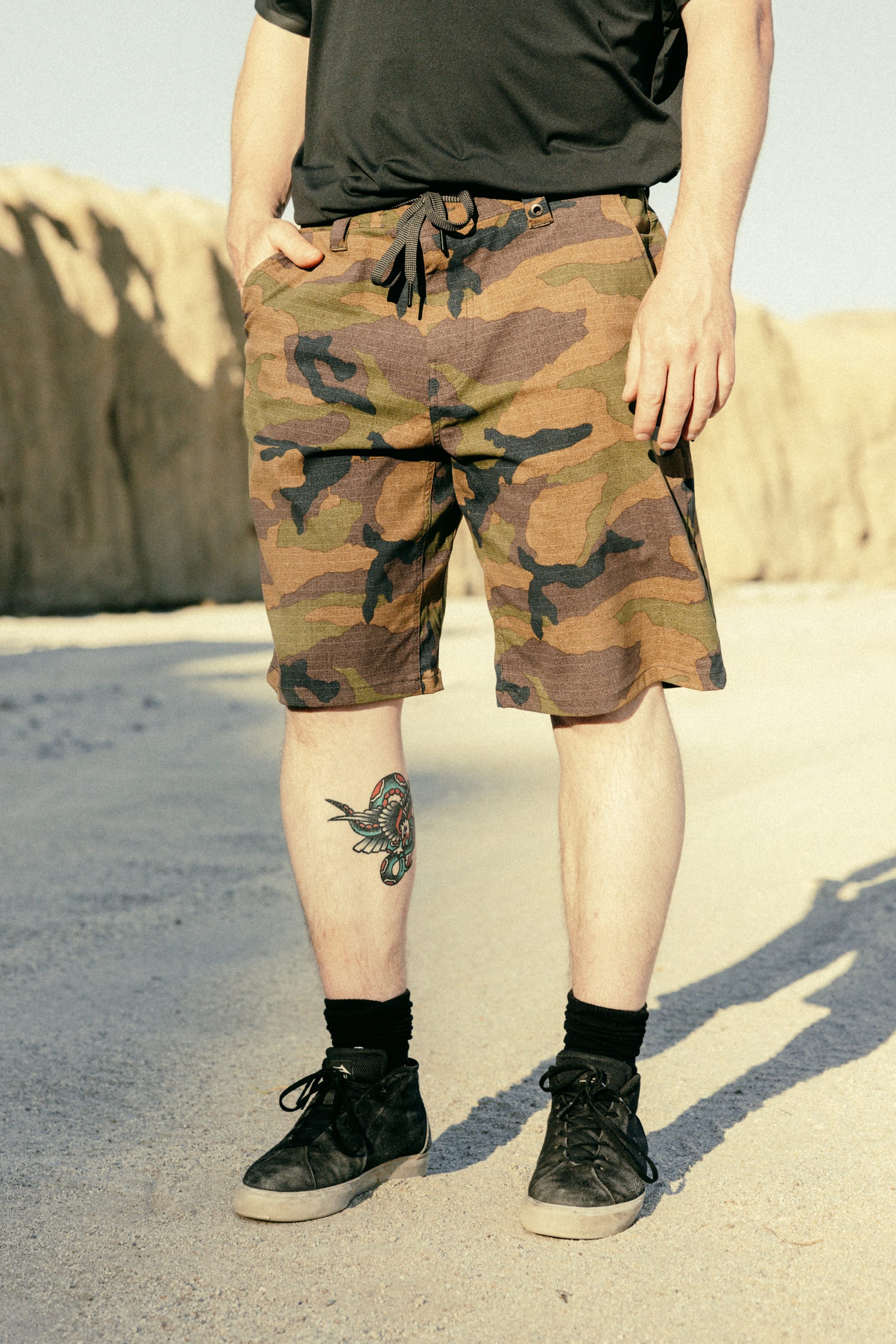 Outdoors on sandy ground, a person wears 686 Men's Everywhere® Hybrid Short - Relaxed Fit with a camo print, a black shirt, black socks, and shoes. A colorful bird tattoo shows on their lower right leg, rocky cliffs in the background.