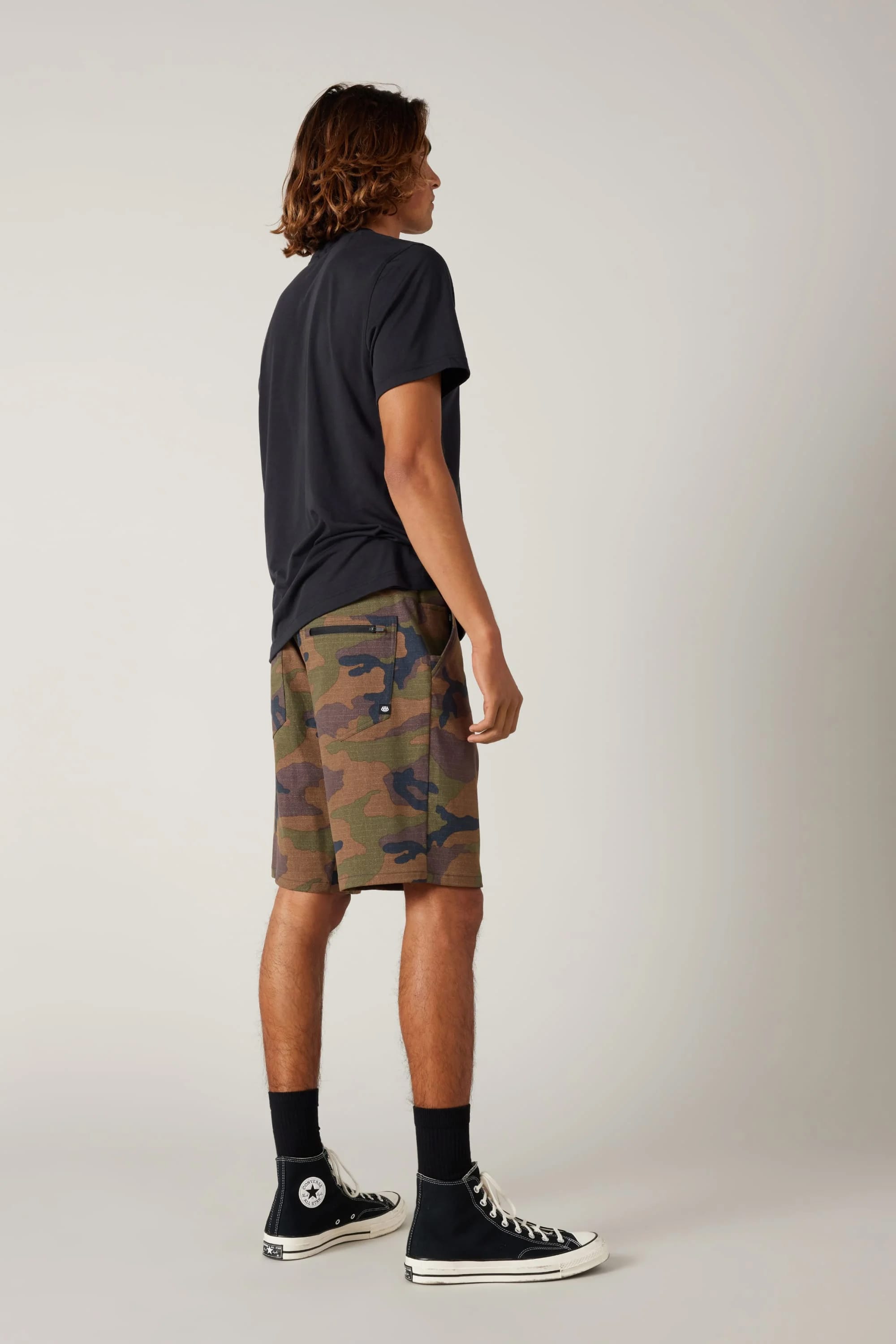 A person stands sideways facing away in a black T-shirt, 686 Men's Everywhere® Hybrid Short - Relaxed Fit by 686, black socks, and black Converse sneakers against a plain light background.