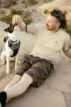 A man wearing sunglasses, a beige shirt, and 686 Men's Everywhere® Hybrid Short - Relaxed Fit sits on a rock feeding a black and white dog in a harness, with rocks and vegetation in the background.