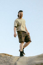 A man, smiling and looking down, stands on a rock under a clear blue sky outdoors wearing beige shorts by 686 Men's Everywhere® Hybrid Short - Relaxed Fit, a beige t-shirt, and black shoes.