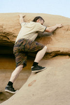 Wearing a beige t-shirt, 686 Men's Everywhere® Hybrid Short - Relaxed Fit, and black sneakers, a person climbs a large light brown rock formation outdoors.