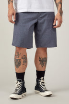 A person wears a white T-shirt and 686 Men's Everywhere® Hybrid Short - Relaxed Fit in blue, paired with black socks and black high-top sneakers. Tattooed arms and legs are visible. The image is cropped at the shoulders, hiding the face.
