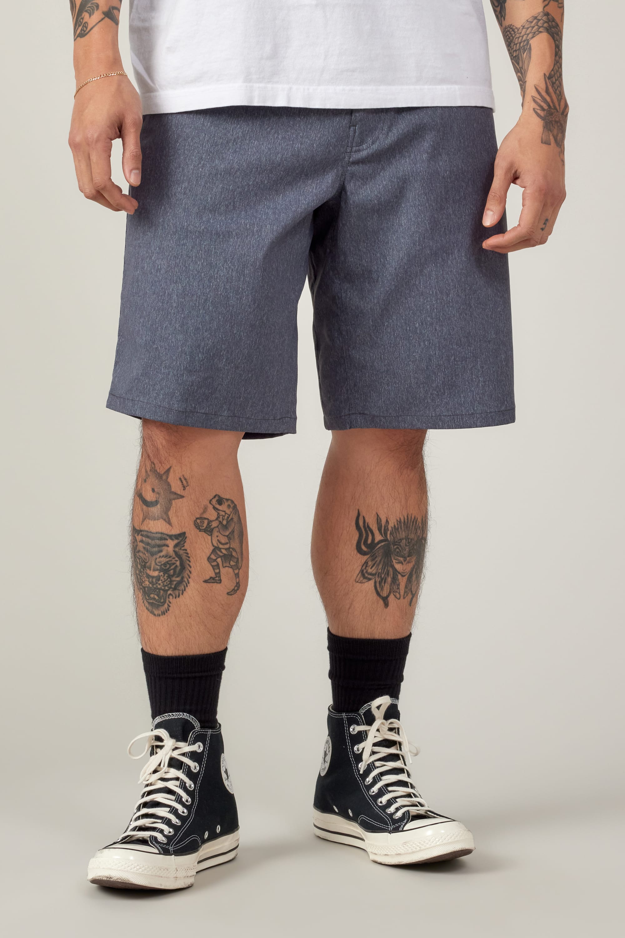 A person wears a white T-shirt and 686 Men's Everywhere® Hybrid Short - Relaxed Fit in blue, paired with black socks and black high-top sneakers. Tattooed arms and legs are visible. The image is cropped at the shoulders, hiding the face.