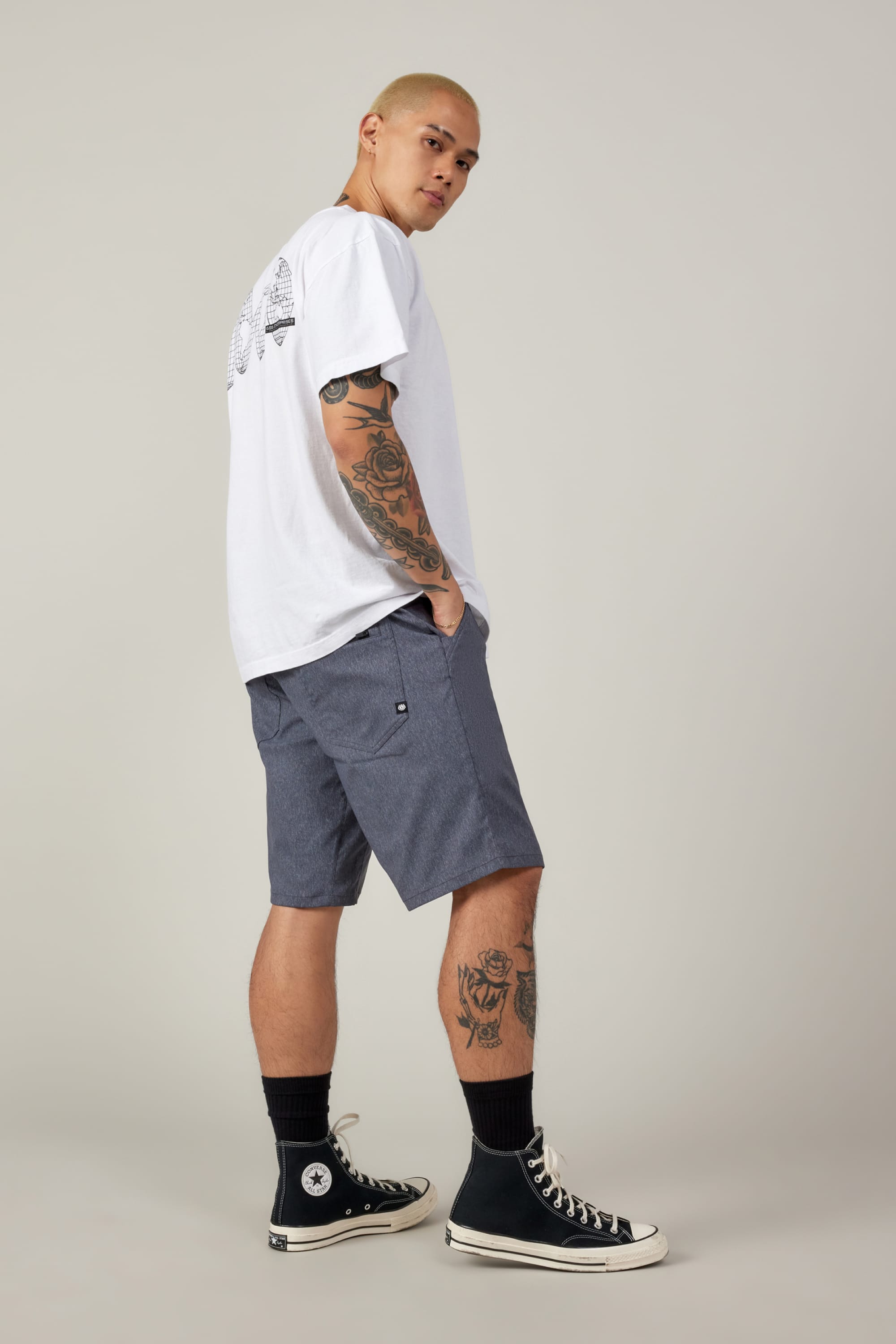 A tattooed person stands sideways, hands in pockets, wearing a white T-shirt, black socks, black high-top sneakers, and 686 Men's Everywhere® Hybrid Short - Relaxed Fit by 686, looking over their shoulder against a neutral background.