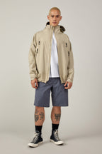 A person with a shaved head wears a beige hooded jacket, white t-shirt, black socks, black Converse, and 686 Men's Everywhere® Hybrid Short - Relaxed Fit by 686. Tattoos are visible on both legs. They stand against a plain light background.