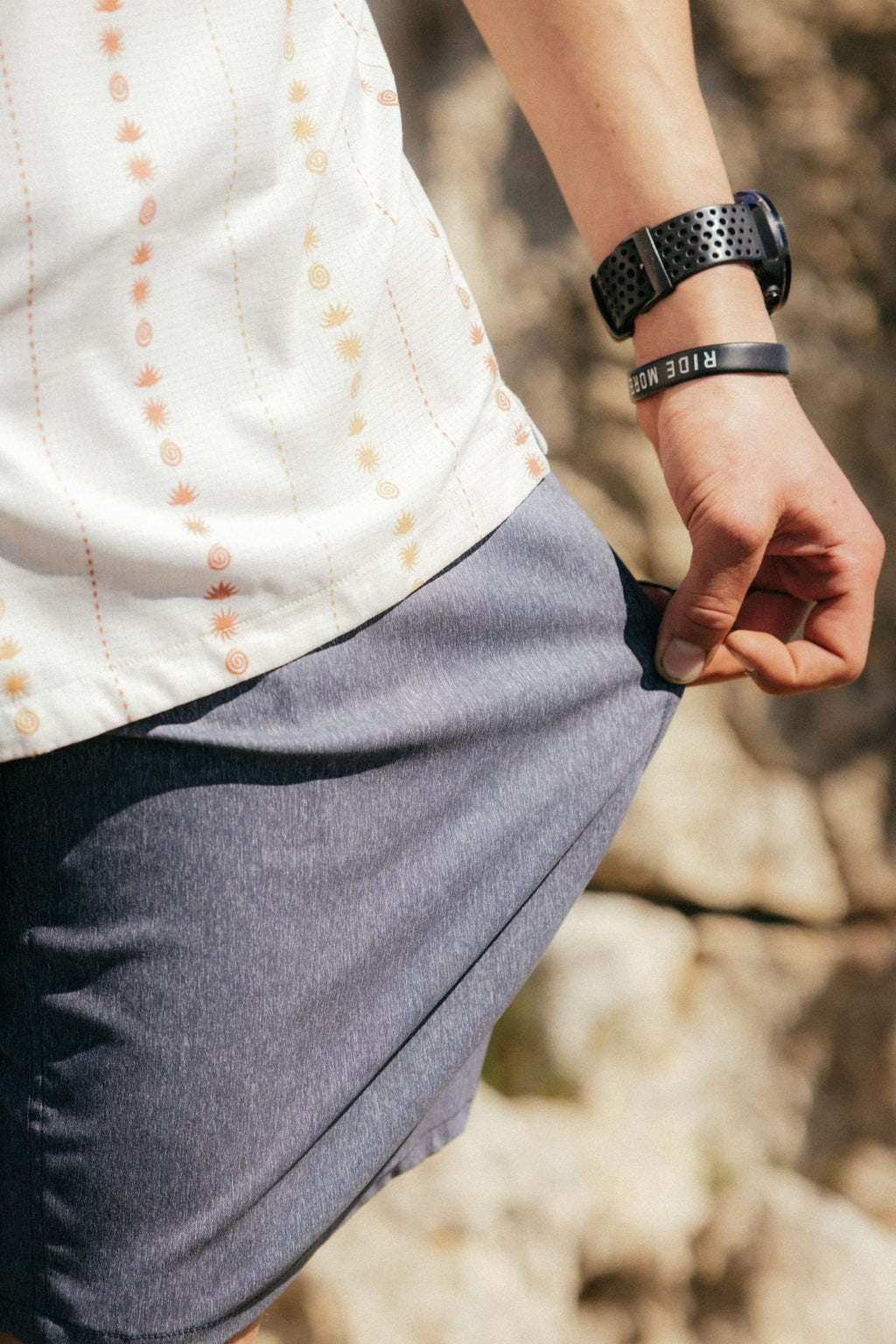 A person in a white patterned shirt wears the 686 Men's Everywhere® Hybrid Short - Relaxed Fit by 686. They hold the hem of their shorts, wearing a black RIDE MORE wristband and watch. Rocks appear in the background.