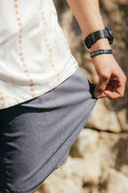 A person in a white patterned shirt wears the 686 Men's Everywhere® Hybrid Short - Relaxed Fit by 686. They hold the hem of their shorts, wearing a black RIDE MORE wristband and watch. Rocks appear in the background.