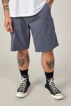 A person stands with hands in pockets, sporting a white tee, 686 Men's Everywhere® Hybrid Short - Relaxed Fit in blue-gray, black crew socks, and black high-top sneakers. Tattoos on their forearms and legs add an edgy touch to the casual vibe.