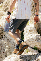 Wearing 686 Men's Everywhere® Hybrid Shorts - Relaxed Fit, dark hiking boots, and a patterned short-sleeve shirt, a tattooed individual climbs rocky terrain with a large water bottle and sports a black watch.