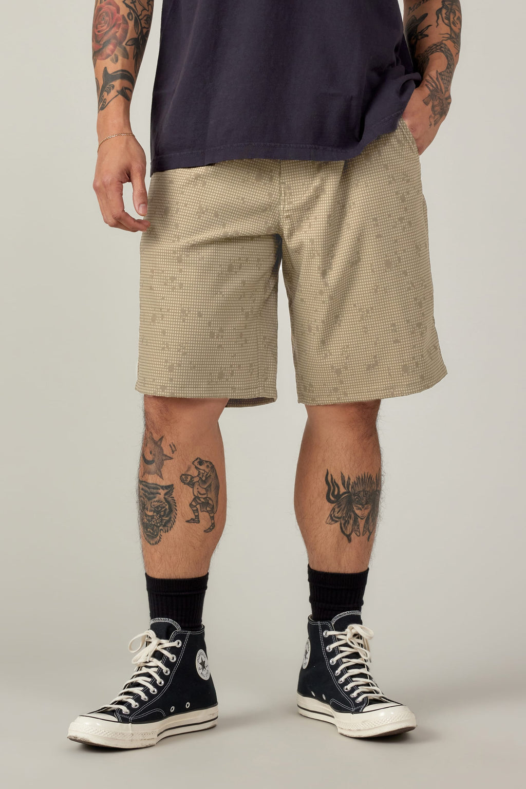 A person wears a dark t-shirt, 686 Men's Everywhere® Hybrid Short - Relaxed Fit in beige check, black socks, and black Converse sneakers. Their tattooed arms and legs stand out against a plain background.
