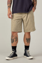 A person wears a dark t-shirt, 686 Men's Everywhere® Hybrid Short - Relaxed Fit in beige check, black socks, and black Converse sneakers. Their tattooed arms and legs stand out against a plain background.