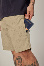 A tattooed person places a passport into the pocket of their 686 Men's Everywhere® Hybrid Short - Relaxed Fit. The beige, patterned shorts are paired with a dark tee, set against a plain background.