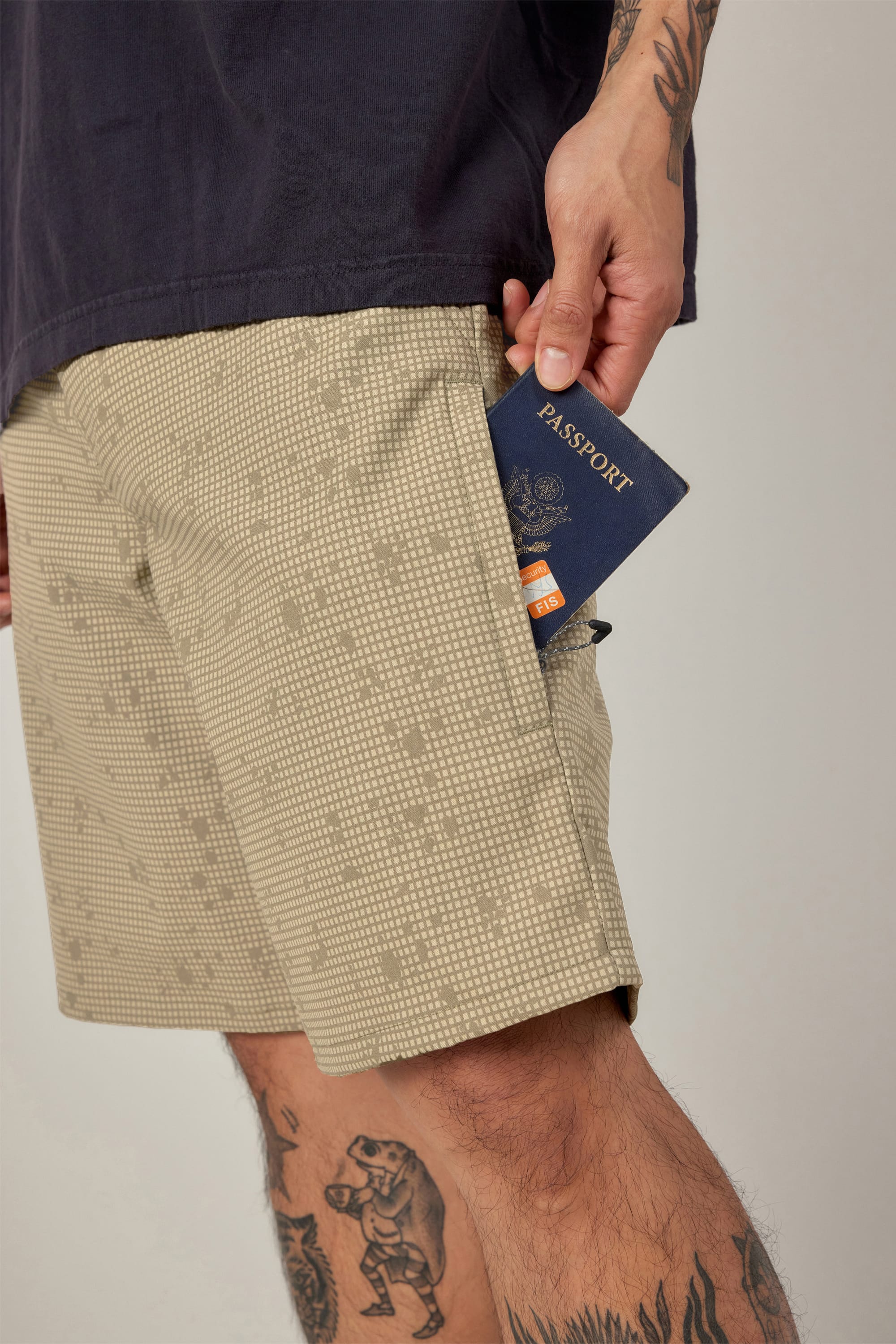 A tattooed person places a passport into the pocket of their 686 Men's Everywhere® Hybrid Short - Relaxed Fit. The beige, patterned shorts are paired with a dark tee, set against a plain background.