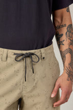 A person wearing 686 Men's Everywhere® Hybrid Short - Relaxed Fit in light beige with a black and white drawstring, paired with a dark shirt. Their tattooed right arm displays "Mama" and a woman's face among other ink designs.