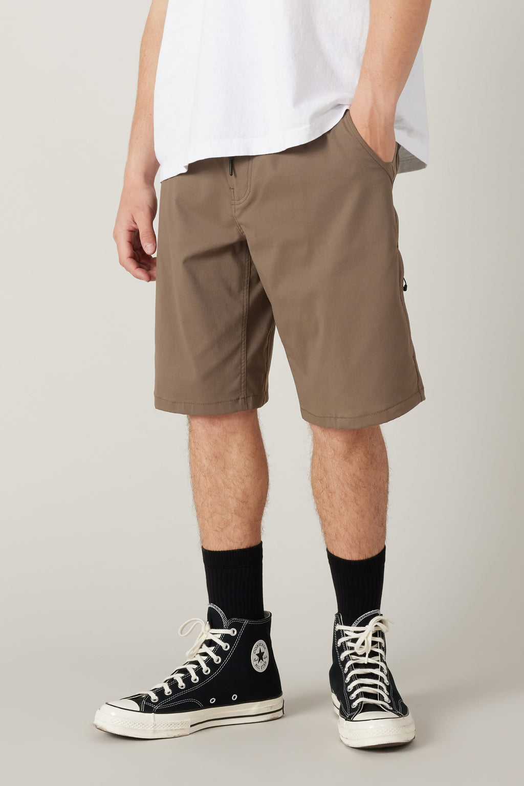 A person wears the 686 Men's Everywhere® Hybrid Short - Relaxed Fit (brown), a white t-shirt, black crew socks, and black Converse high-tops, standing with one hand in their pocket against a plain background.