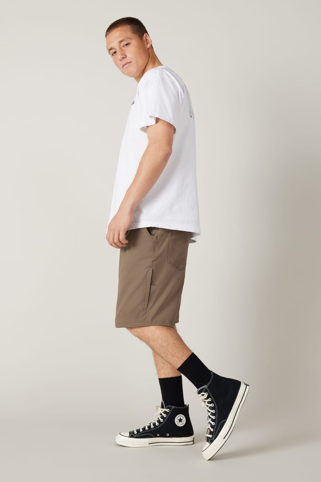 A young man stands sideways, looking back at the camera. He wears a white T-shirt and 686 Men's Everywhere® Hybrid Short - Relaxed Fit in brown, paired with black socks and high-top sneakers against a plain, light background.