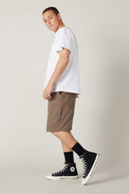 A young man stands sideways, looking back at the camera. He wears a white T-shirt and 686 Men's Everywhere® Hybrid Short - Relaxed Fit in brown, paired with black socks and high-top sneakers against a plain, light background.