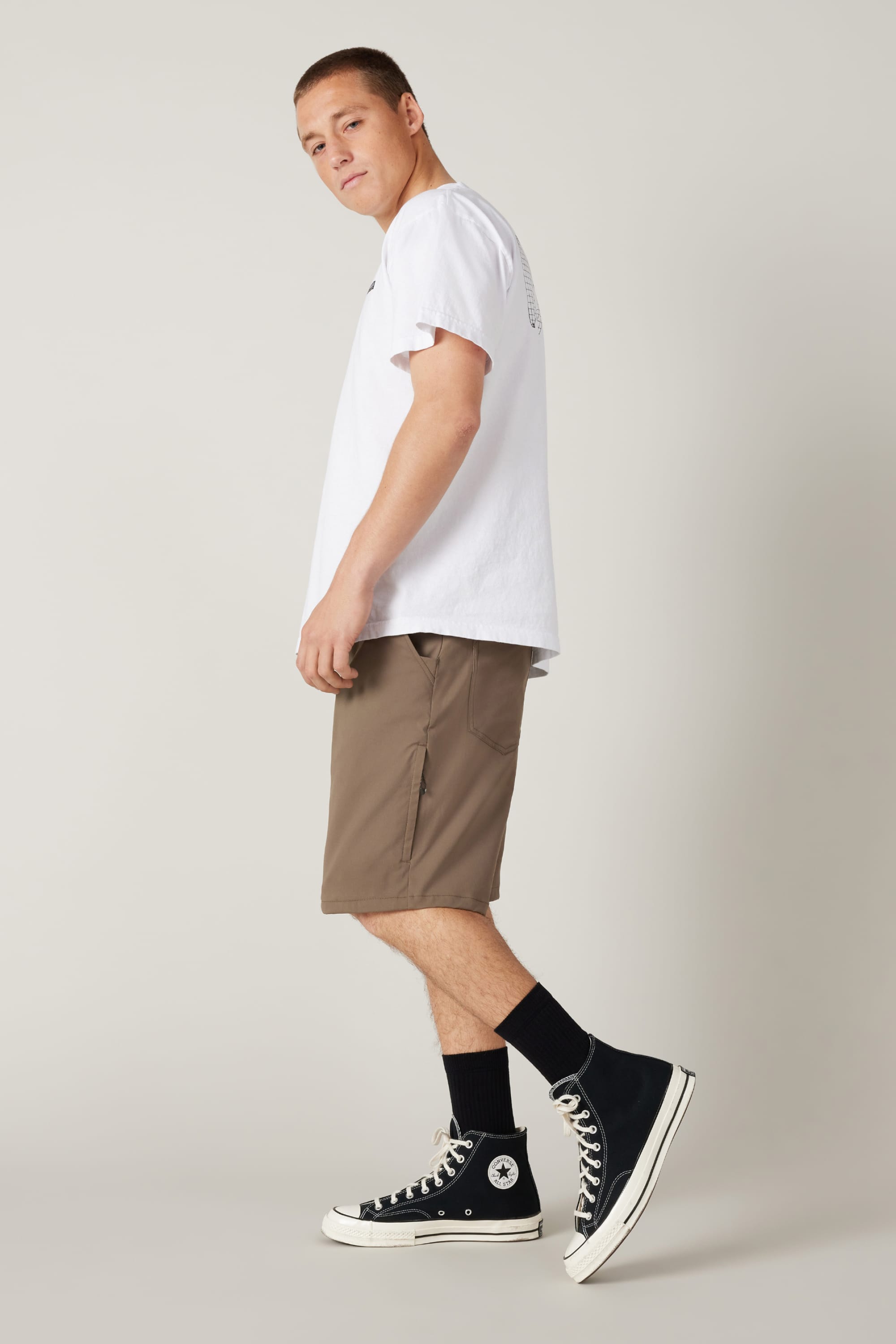 A young man stands sideways, looking back at the camera. He wears a white T-shirt and 686 Men's Everywhere® Hybrid Short - Relaxed Fit in brown, paired with black socks and high-top sneakers against a plain, light background.