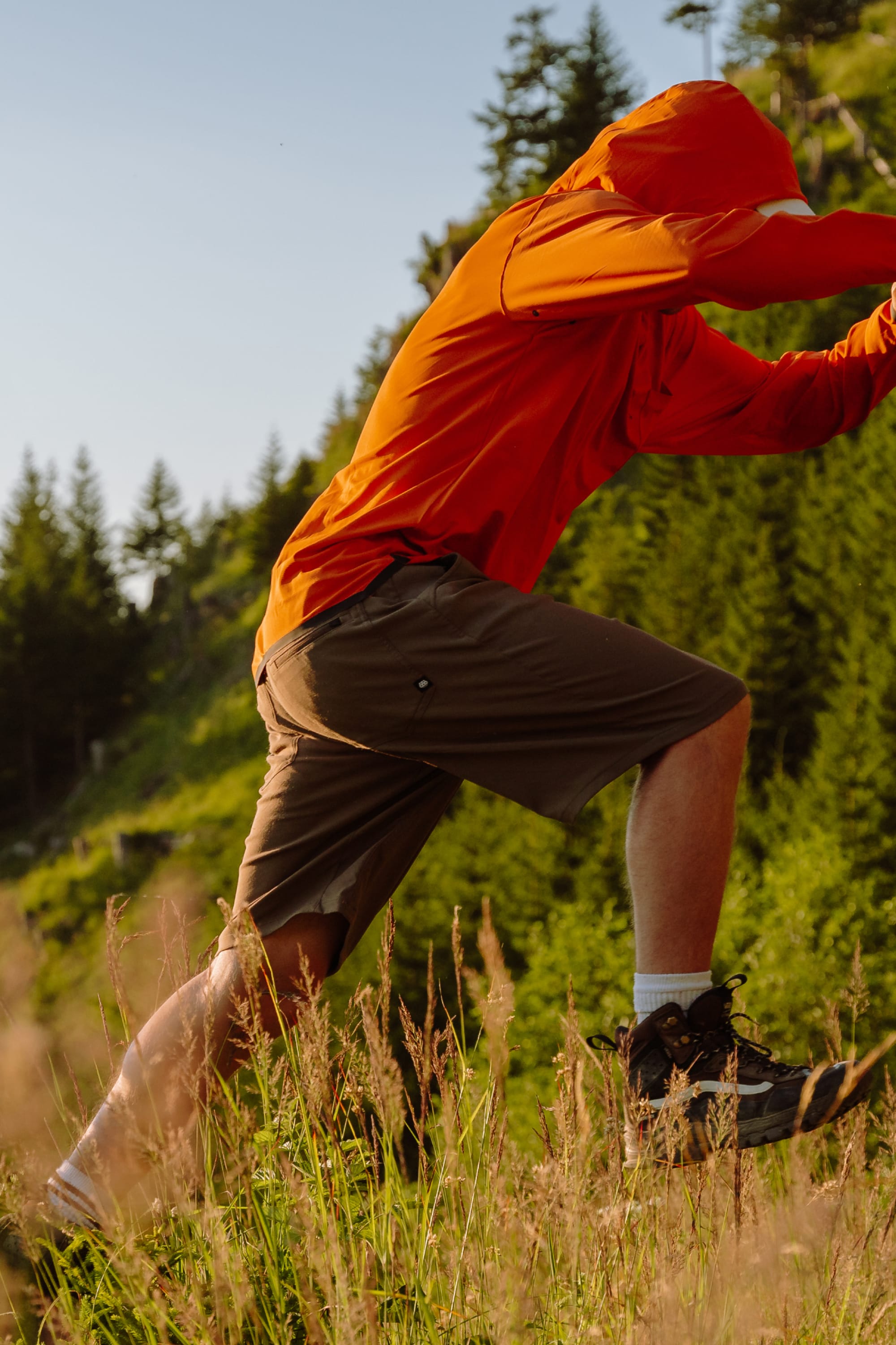 Wearing 686 Men's Everywhere® Hybrid Short - Relaxed Fit, a person in an orange jacket hikes uphill through tall grass, with green trees and a blue sky behind; their face is hidden.