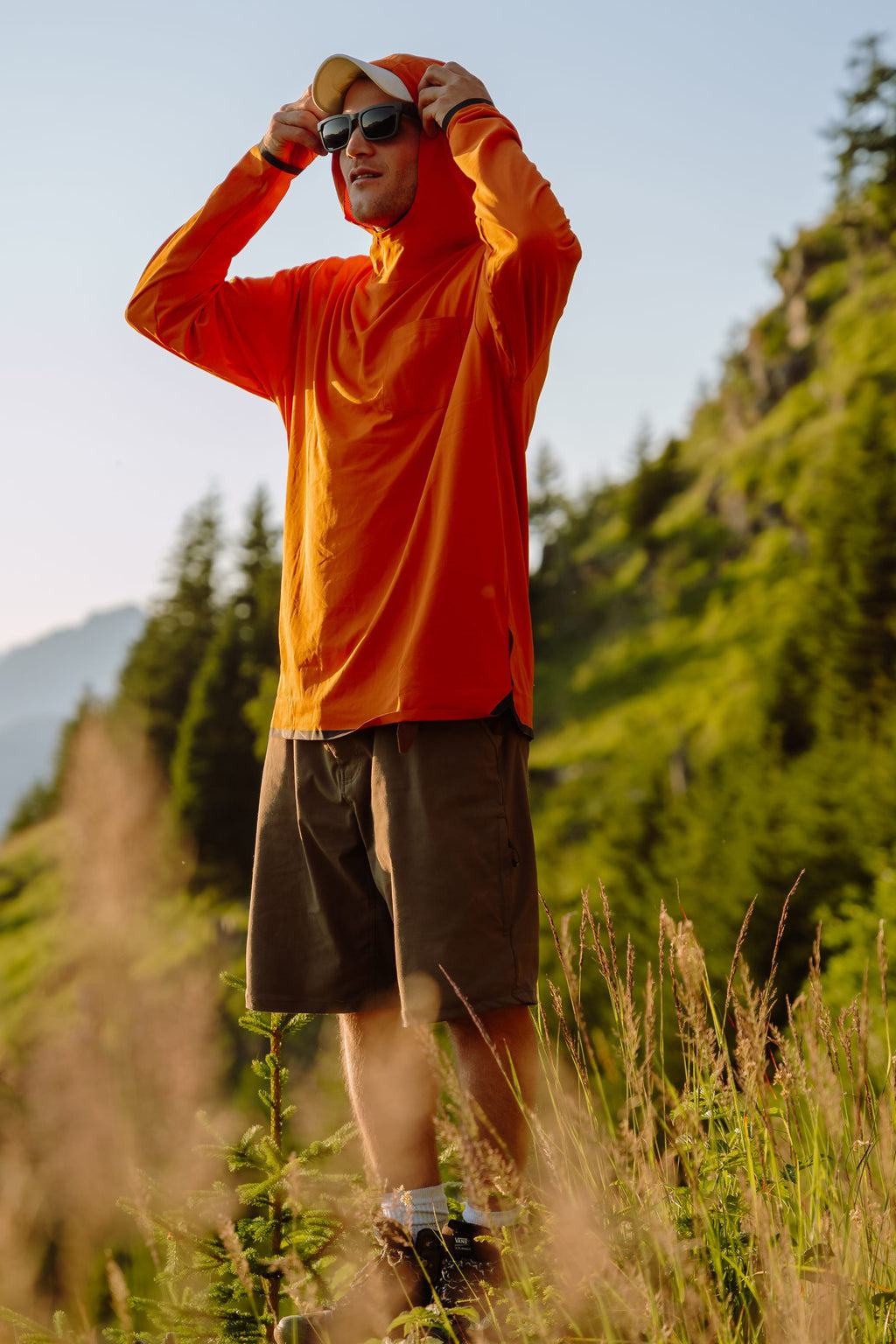 A man in an orange hoodie, sunglasses, and a cap stands on grass wearing 686 Men's Everywhere® Hybrid Short - Relaxed Fit, with a mountain landscape and pine trees behind him on a sunny day.