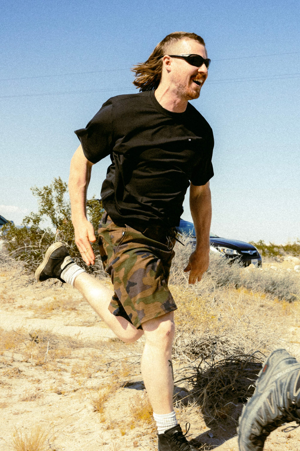 Wearing sunglasses and the 686 Men's Let's Go Tech Tee, a man runs and smiles outdoors through a sunny, desert-like landscape with dry bushes and blue sky—ideal for spring or summer adventure from 686.