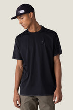 A man in the 686 Men's Let's Go Tech Tee—plain black with a small chest logo—and olive green pants stands against a light background, looking down to his left, ready for any spring or summer adventure.