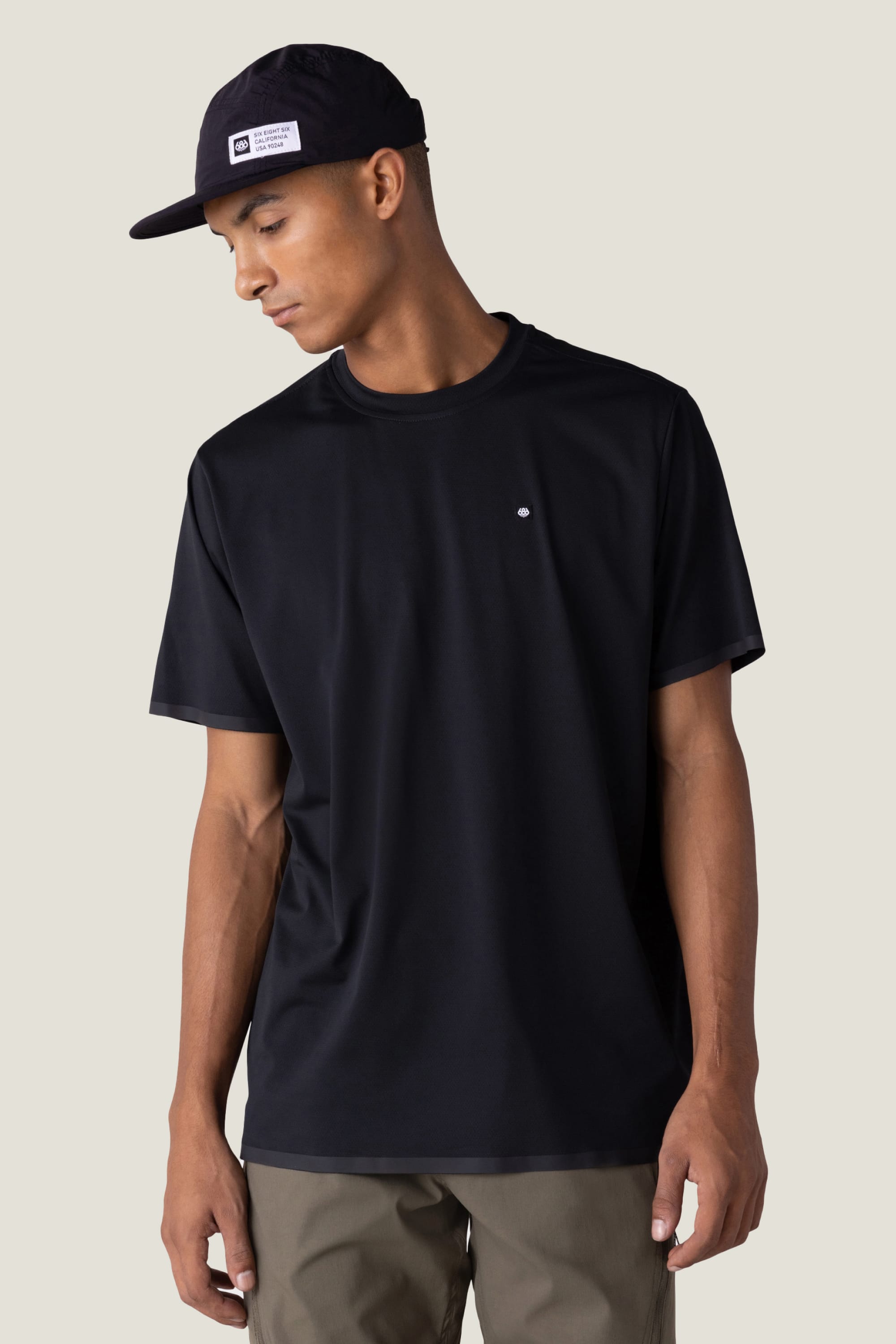 A man in the 686 Men's Let's Go Tech Tee—plain black with a small chest logo—and olive green pants stands against a light background, looking down to his left, ready for any spring or summer adventure.