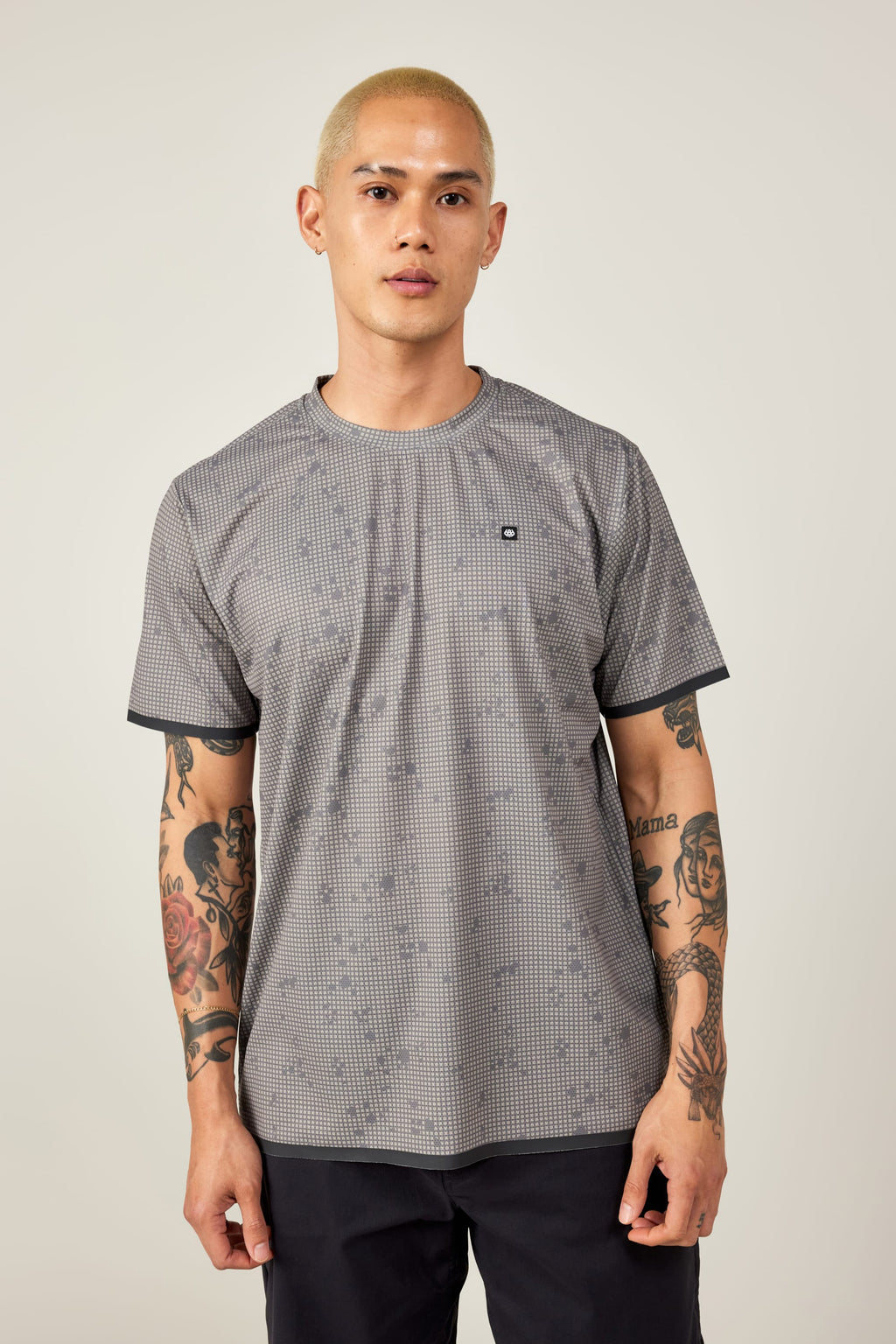A person with short blond hair and arm tattoos wears the 686 Men's Let's Go Tech Tee by 686, featuring a gray pattern, paired with black pants, standing against a plain light background.