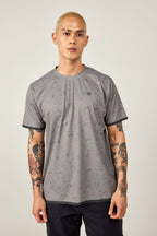 A person with short blond hair and arm tattoos wears the 686 Men's Let's Go Tech Tee by 686, featuring a gray pattern, paired with black pants, standing against a plain light background.
