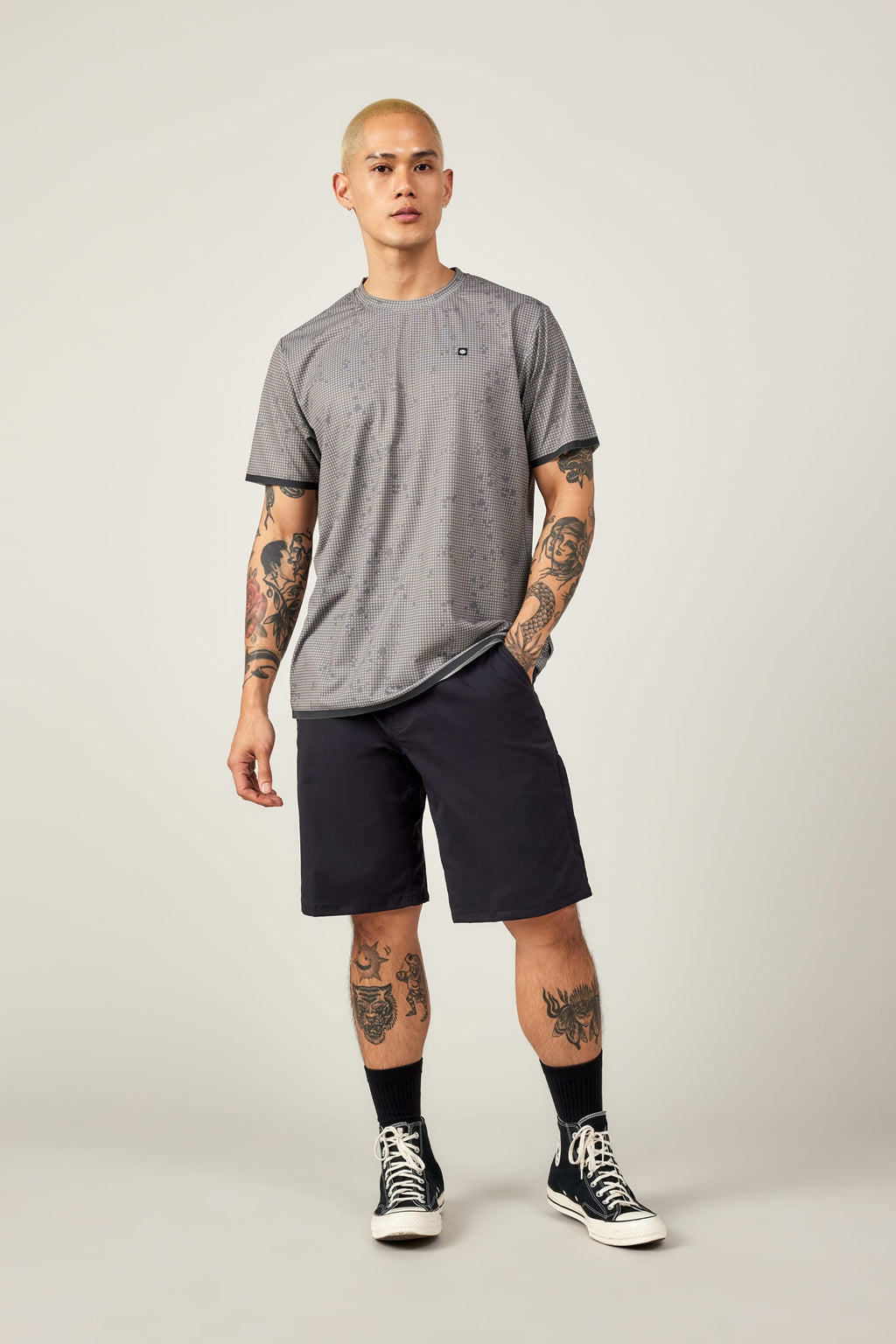 A man with short blond hair and tattoos wears the 686 Men's Let's Go Tech Tee, black shorts, socks, and sneakers, standing against an off-white background—perfectly outfitted by 686 for spring-summer adventures.