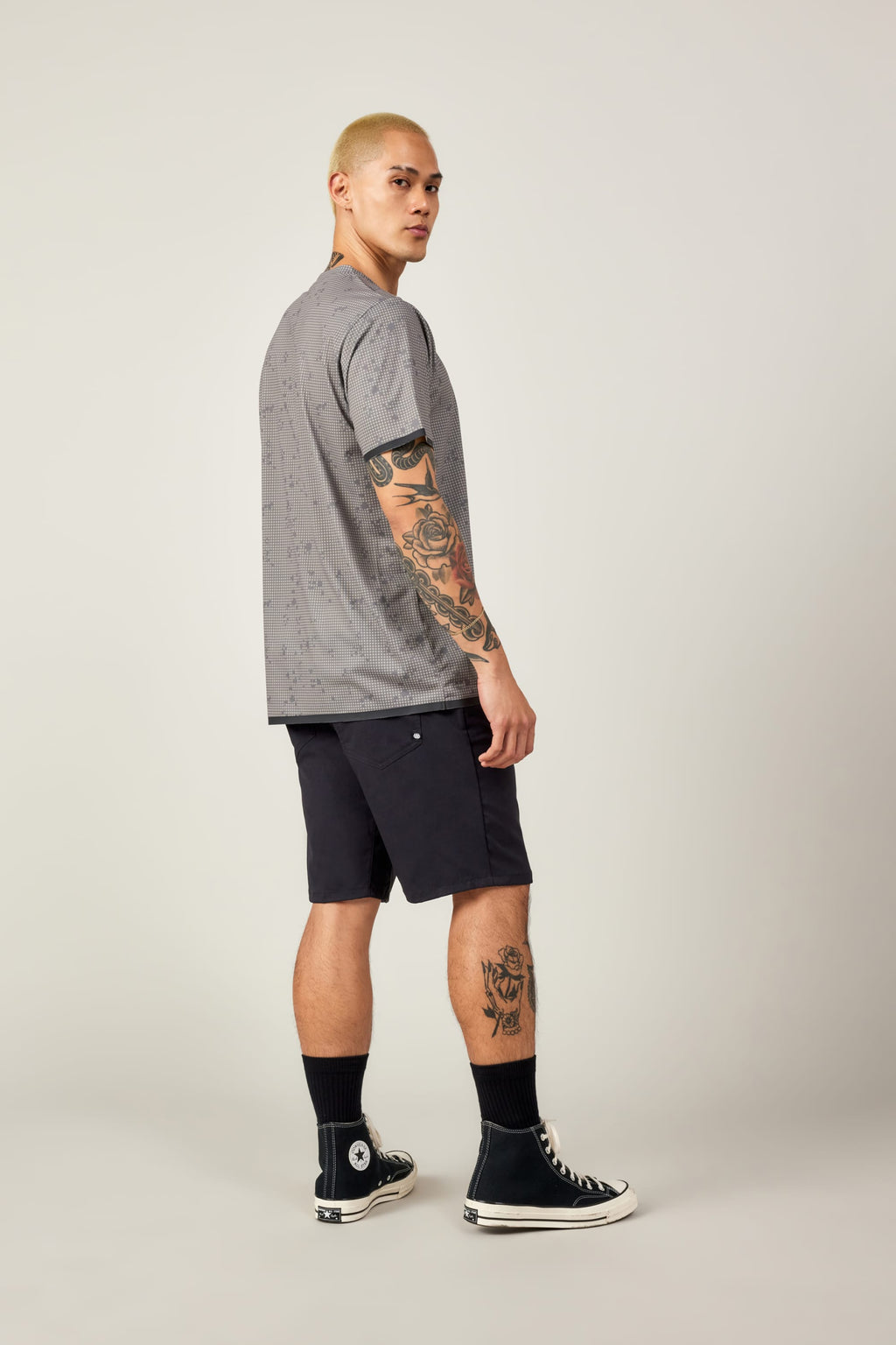 A person with short blond hair and visible tattoos wears the 686 Men's Let's Go Tech Tee by 686 in gray, paired with black shorts, black crew socks, and black Converse sneakers, standing slightly turned to the side against a plain background.