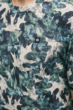 The 686 Men's Let's Go Tech Tee is a lightweight shirt with a green, white, and dark blue leafy camo pattern and a small black patch with a white geometric symbol—ideal for spring and summer adventures.