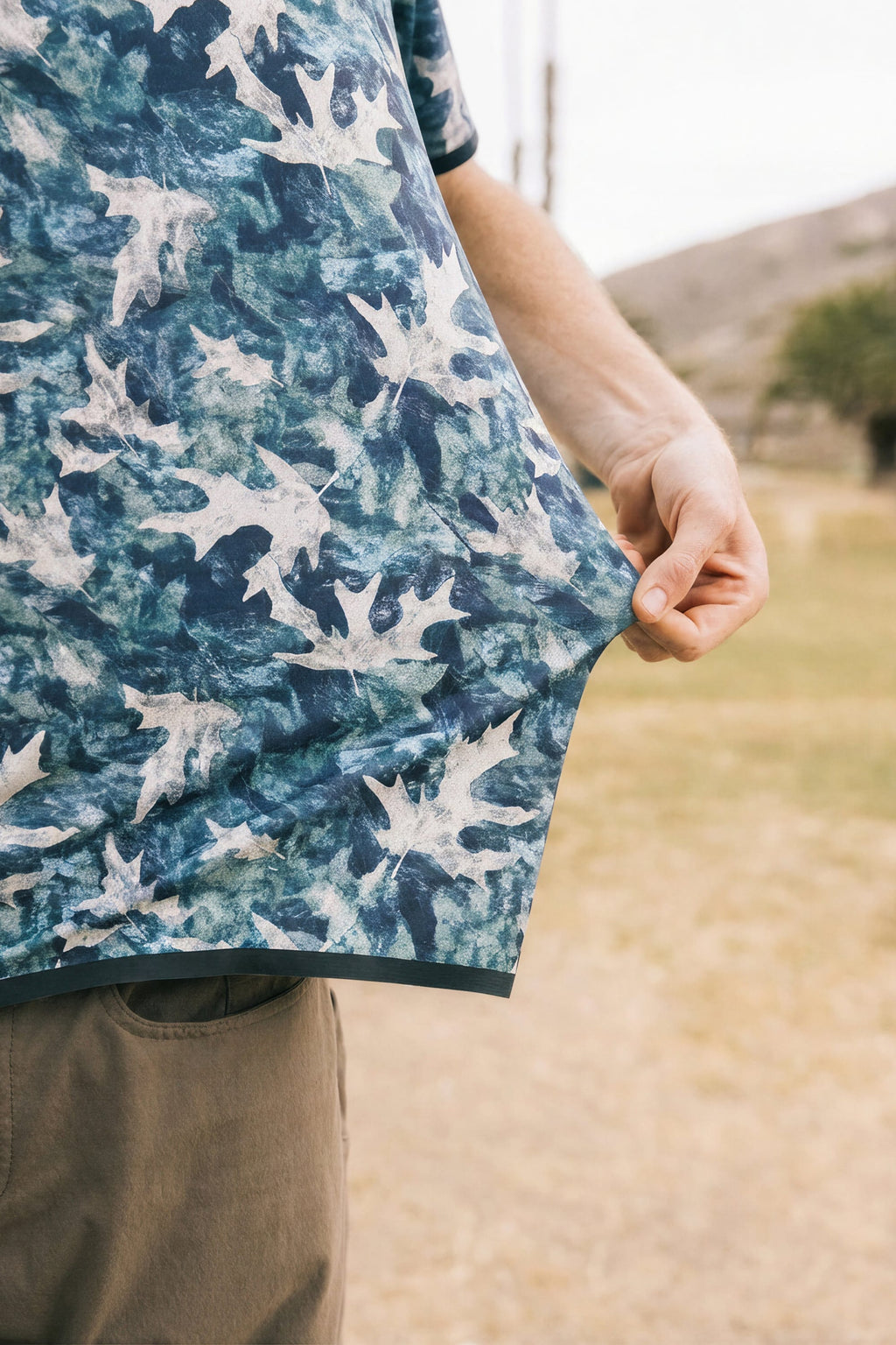 A man in the 686 Men's Let's Go Tech Tee, a blue breathable shirt with a white leaf print, stands outdoors on grass—ideal spring-summer attire for adventures among hills and trees.