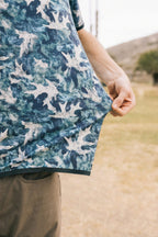 A man in the 686 Men's Let's Go Tech Tee, a blue breathable shirt with a white leaf print, stands outdoors on grass—ideal spring-summer attire for adventures among hills and trees.