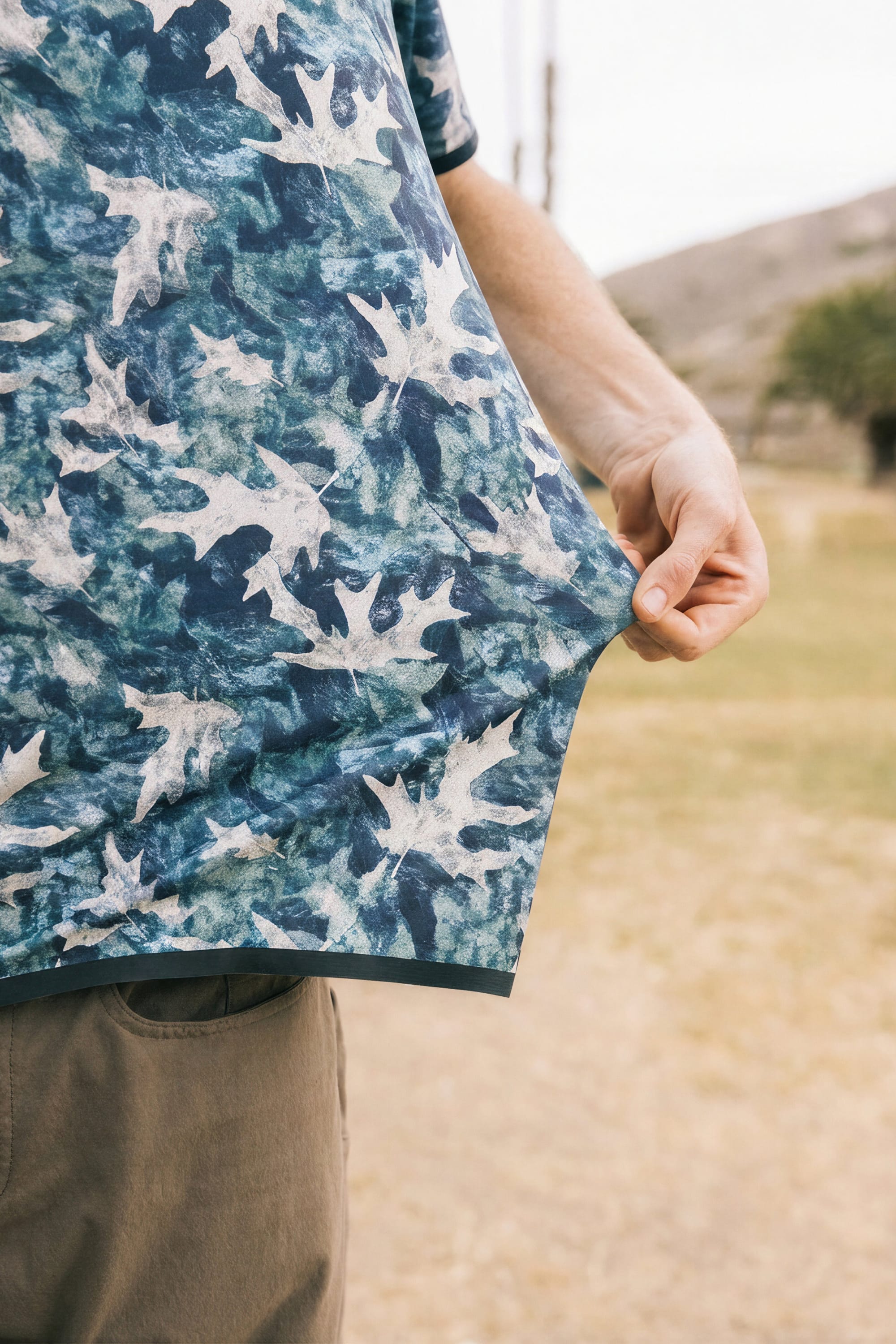 A man in the 686 Men's Let's Go Tech Tee, a blue breathable shirt with a white leaf print, stands outdoors on grass—ideal spring-summer attire for adventures among hills and trees.